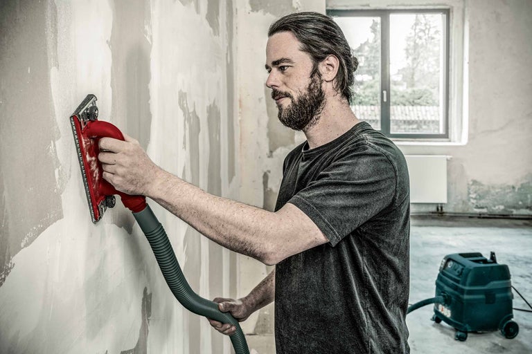 Man schuurt muur met een gipsplaatschuurmachine, aangesloten op een stofzuiger