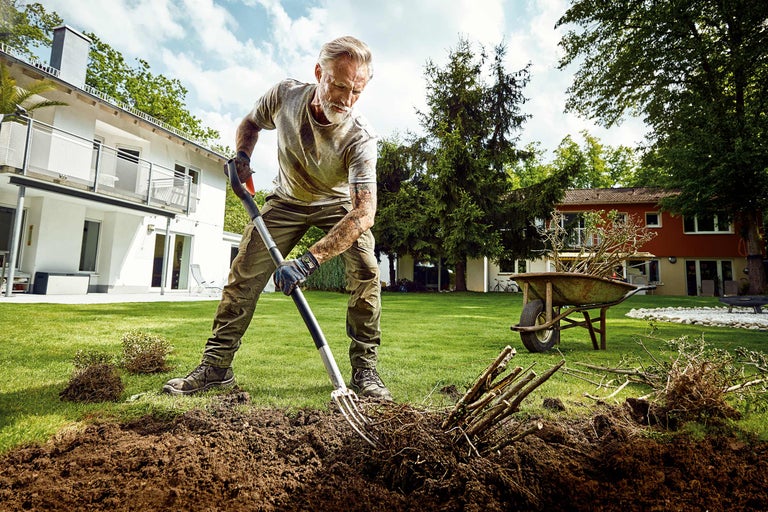 Een man werkt met een greep in de tuin, verplante struiken en een kruiwagen op de achtergrond.