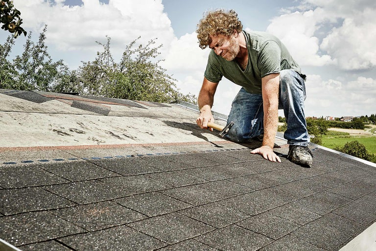 Een man bevestigt bitumineuze dakshingles op een dak met een hamer.