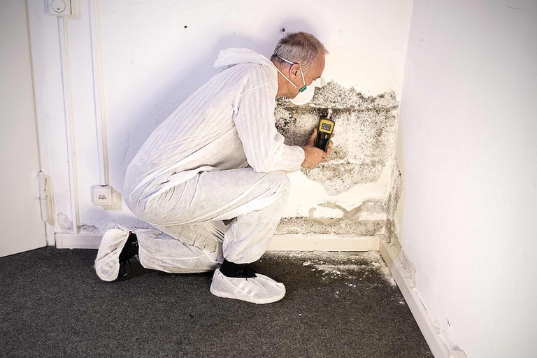 Een man in beschermende kleding inspecteert schimmel op een muur met een meetapparaat.