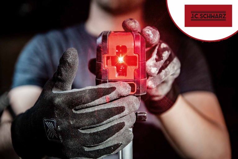 Een persoon houdt een kruislijnlaser vast met handschoenen aan. J.C. Schwarz logo.