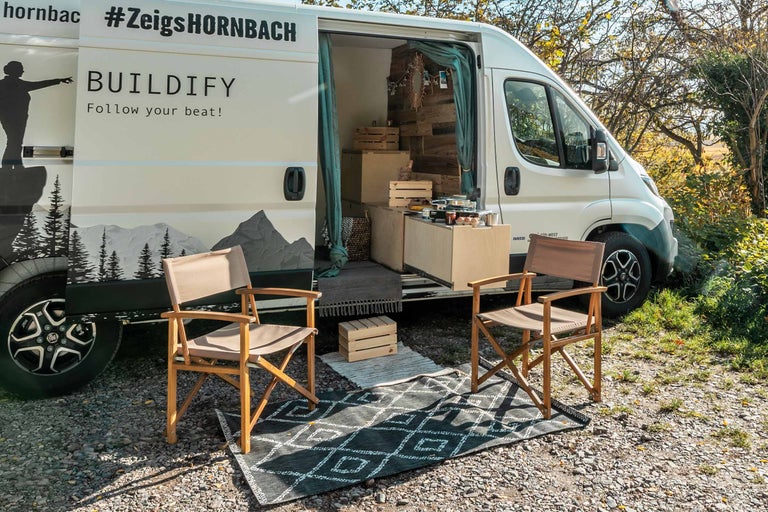 Scene met een witte bus, met twee houten stoelen op een vloerkleed met patroon ervoor
