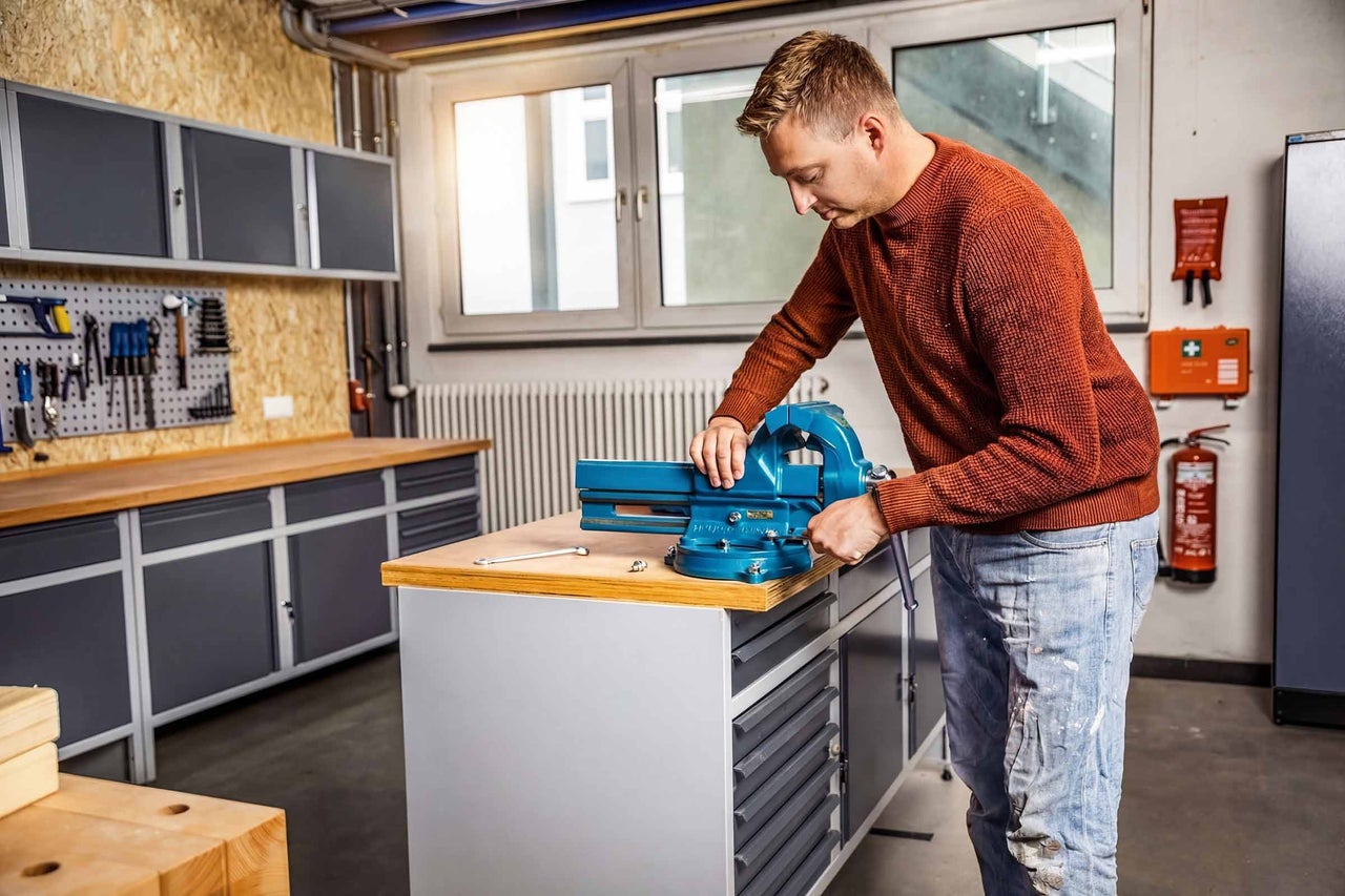 Man monteert een blauwe bankschroef op een werkbank in een lichte, georganiseerde werkplaats met gereedschapswand en wandkasten.