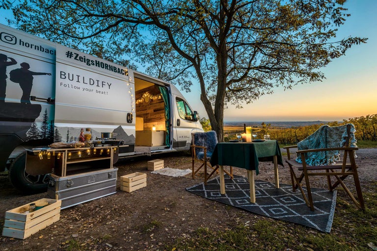 Scène met kampeerbus, tafel, stoelen en kookgerei onder een boom bij zonsondergang