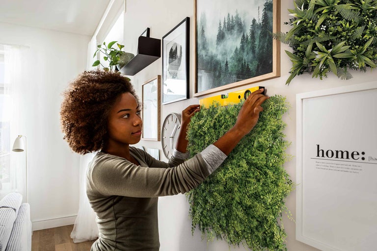 Vrouw monteert een groene plantenwand met een waterpas aan een muur, omringd door decoraties. Lichte, georganiseerde ruimtesfeer.