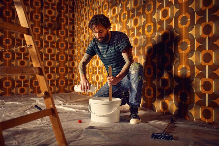 Man mengt verf in een emmer voor renovatiewerkzaamheden in een kamer met retro behang.