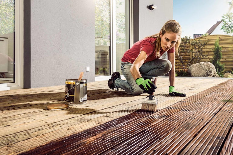 Vrouw schildert een houten terras met een kwast en een blik houtbeits.