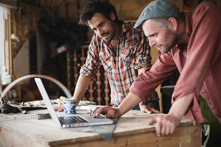 Twee ambachtslieden werken aan een laptop in een werkplaats. Gereedschap en bouwmaterialen zijn op de achtergrond te zien.