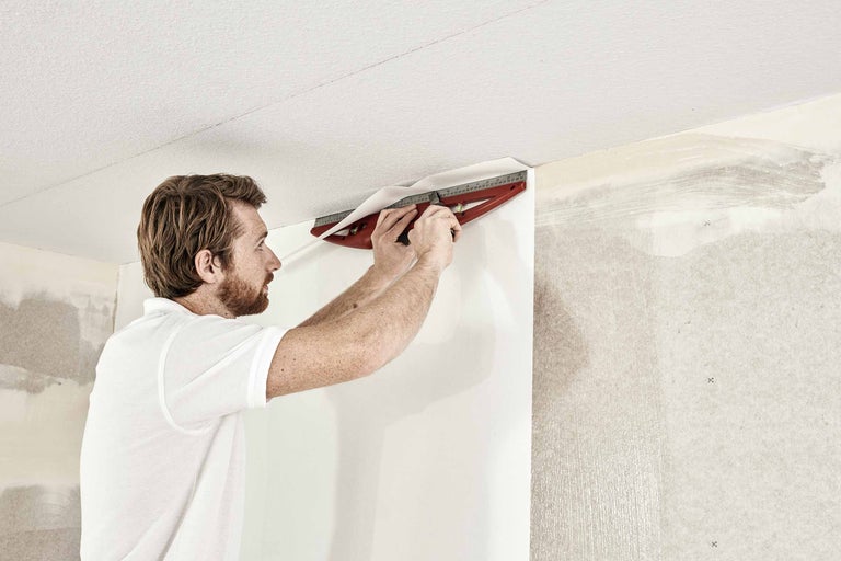 Een man snijdt behang met een snijmal op een muur.