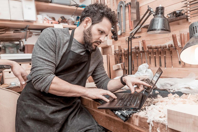 Een vakman met een schort bedient een laptop in een werkplaats.