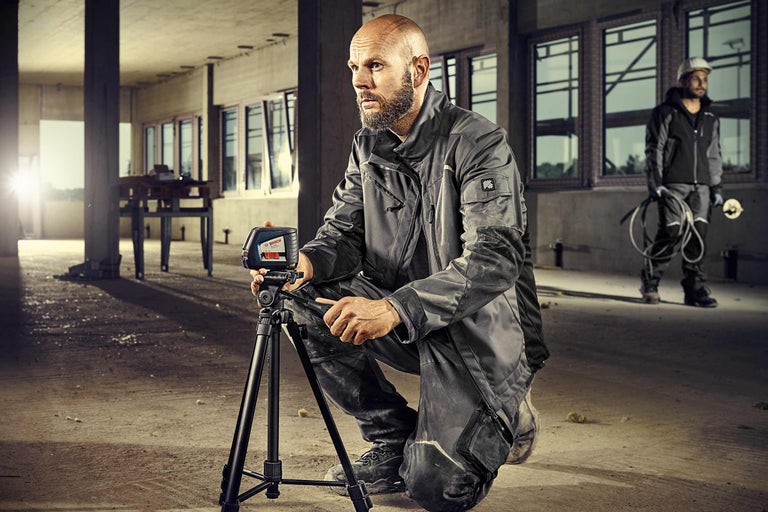 Een werknemer hurkt met een kruislijnlaser en een statief op een bouwplaats.