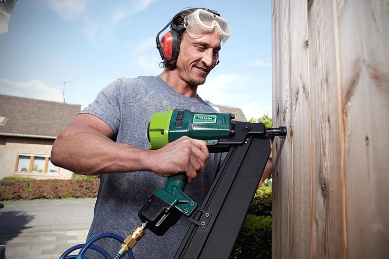 Een man bevestigt een houten plank aan een houten schutting met een pneumatische tacker. De man draagt een veiligheidsbril en gehoorbescherming. Prebena logo.