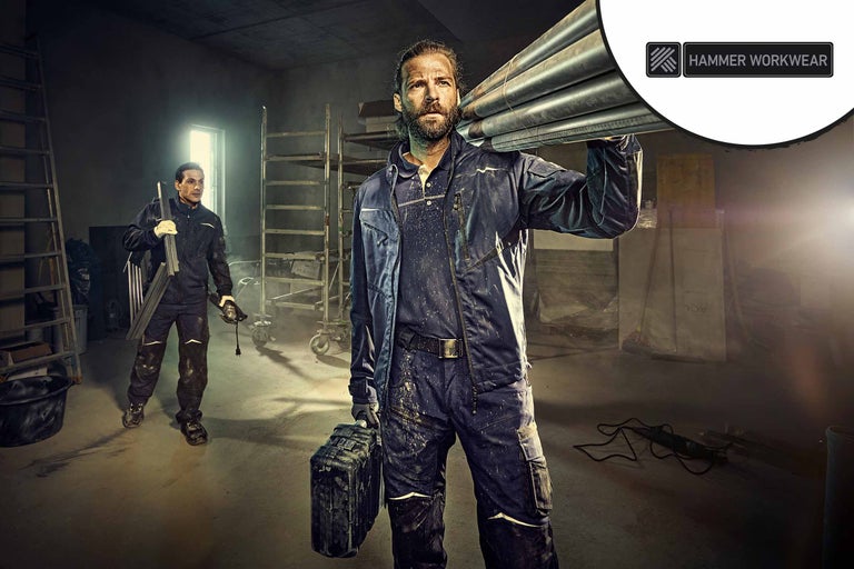 Twee arbeiders in werkkleding in een lichte, georganiseerde ruimte. Eén draagt buizen, de andere een slijpmachine. Hammer Workwear Logo