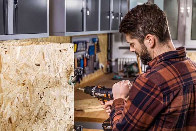 Man gebruikt Worx accuboorschroefmachine op OSB-plaat in werkplaats met wandkasten en gereedschapshouders.