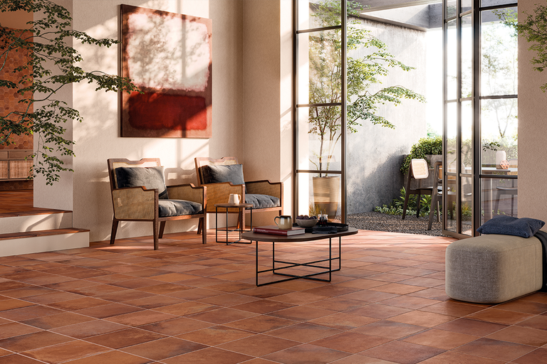 Lichte woonkamer met vierkante vloertegels in terracotta-look, twee houten fauteuils en zicht op de buitenruimte door grote glazen deuren.