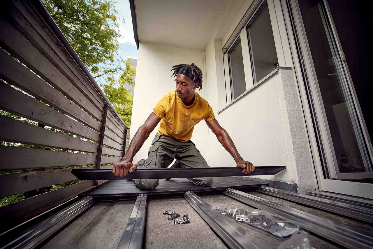 Een man installeert donkere vlonderplanken op een metalen onderbouw op een licht balkon met een houten reling.