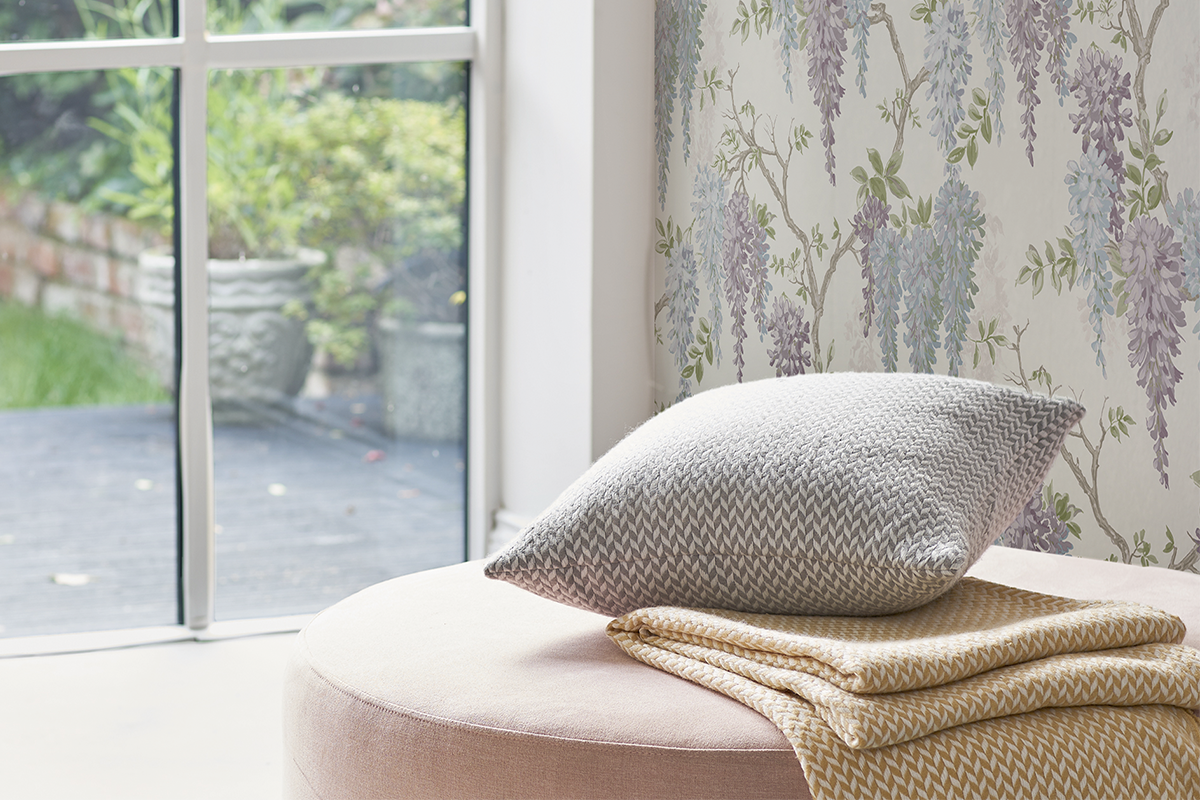 Woonkamer met vliesbehang met blauweregen motief, ronde poef met sierkussen en plaid voor een kamerhoog raam met uitzicht op de tuin