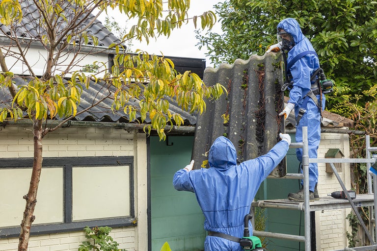Twee personen in beschermende kleding verwijderen een asbestcementplaat van een huis.