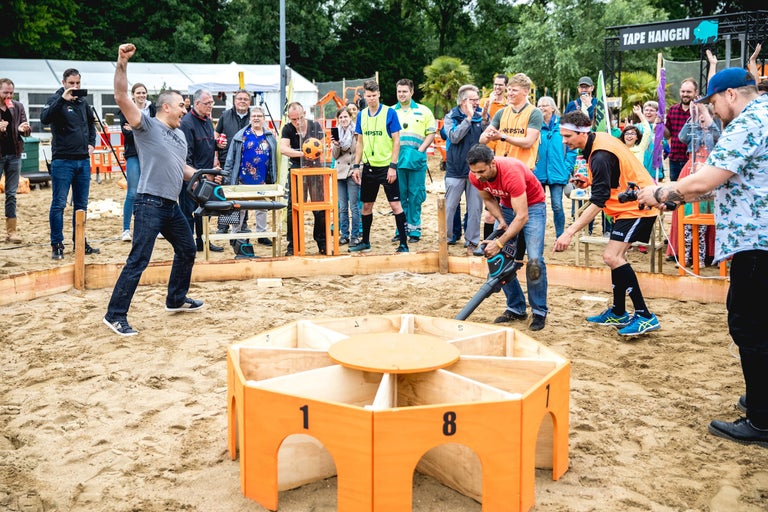 Buitenevenement met deelnemers en toeschouwers rond een zandbak met houten elementen.