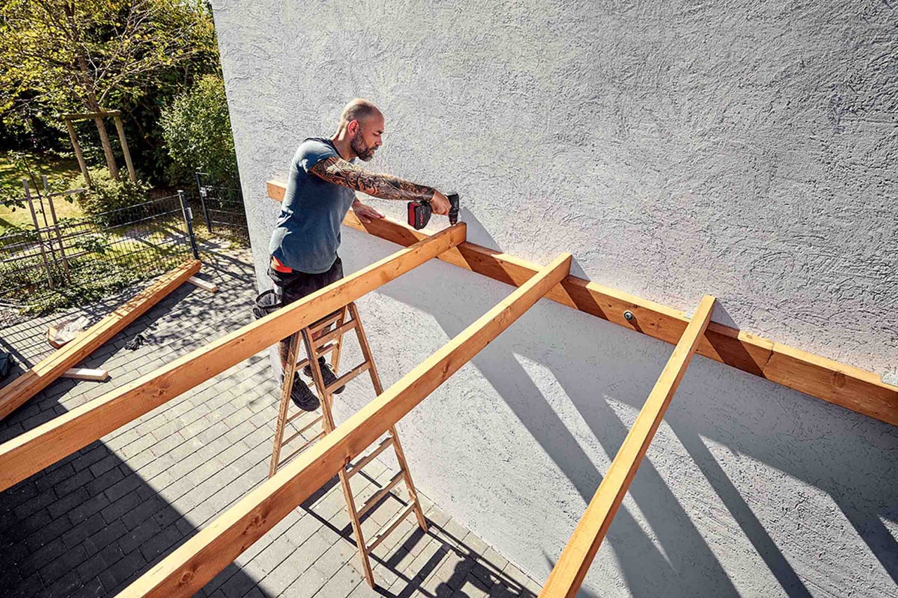 Een man op een houten ladder bevestigt houten balken aan een witte muur met een accuboormachine voor de bouw van een terrasoverkapping.