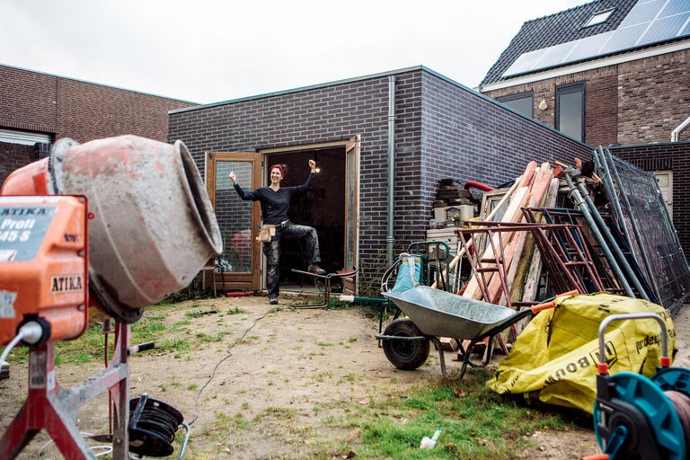 Bouwplaats met betonmolen, kruiwagen, ladder en vrouw in de deuropening