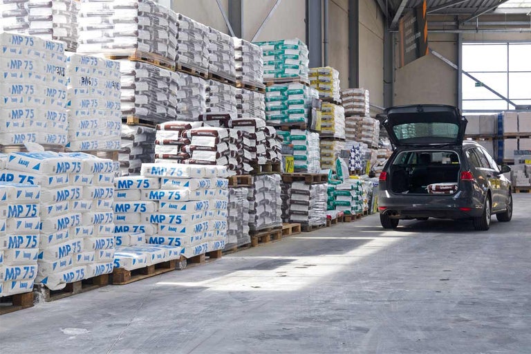 Schappen in een bouwmarkt gevuld met pallets gips en mortel, een auto met open kofferbak staat klaar.