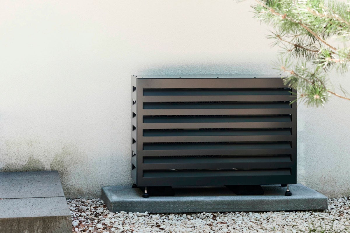 Donkere omkasting voor warmtepomp of airconditioning met lamellen, buiten geplaatst tegen een muur op een betonnen sokkel.