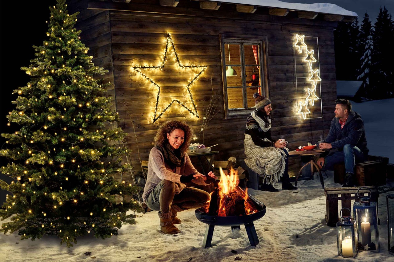 Winterse scène met een vrouw bij een kampvuur, een verlichte kerstboom en stervormige verlichting op een houten hut.