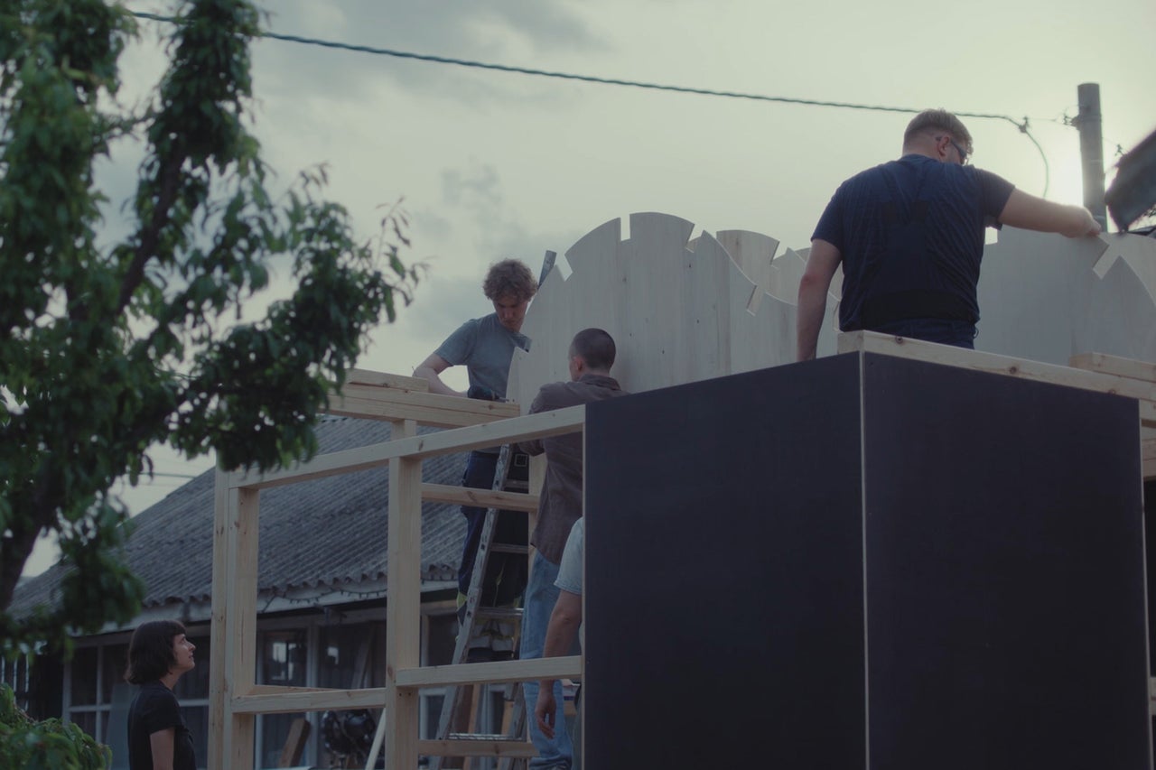 Drie personen monteren overdag een houten dakconstructie met gevormde multiplex platen op een frame van houten balken in de buitenlucht.