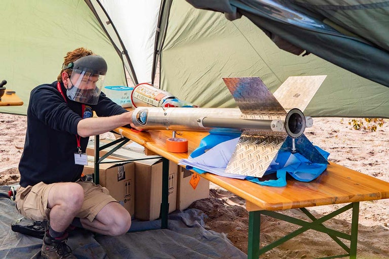 Een persoon met gelaatsbescherming die aan een raketmodel op een tafel werkt.