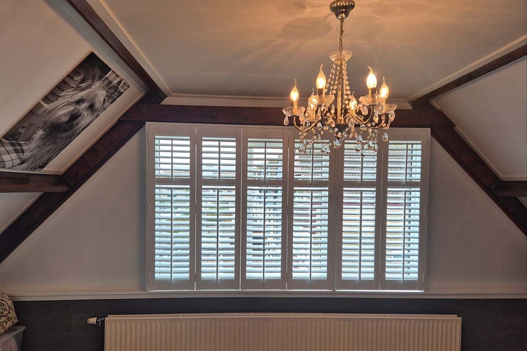 Woonruimte met schuin plafond en witte shutters voor het raam, inclusief een kristallen kroonluchter aan het plafond.