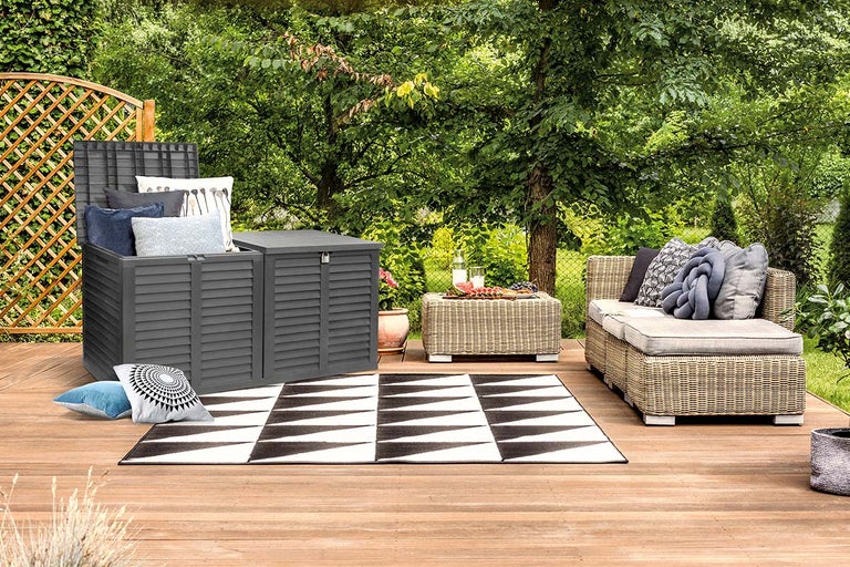 Tuinsetting met grijze opbergbox, rotanmeubels en een geometrisch tapijt op een houten terras met groene achtergrond