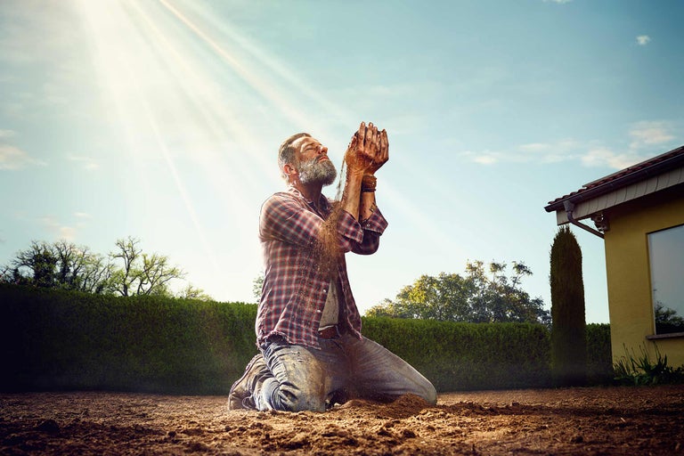 Een man knielt op de grond en laat aarde door zijn handen glippen, met een huis en bomen op de achtergrond.