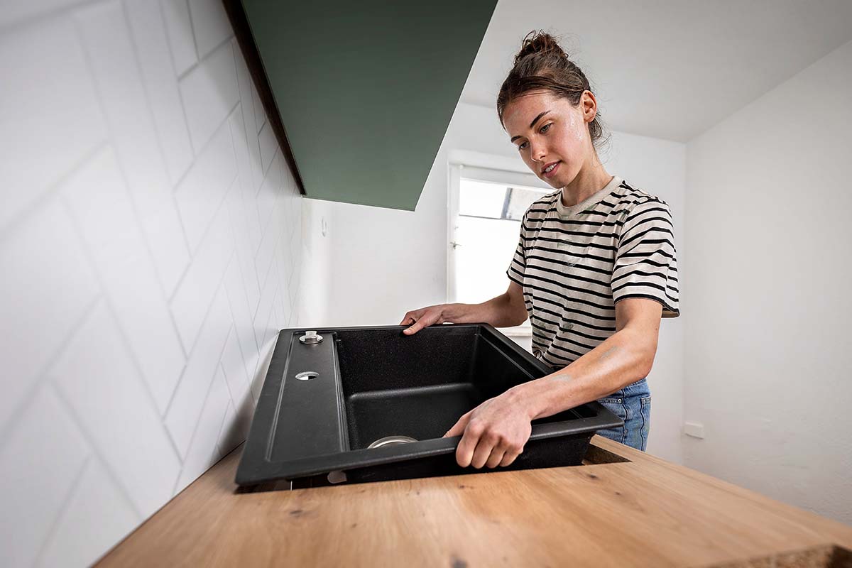 Iemand installeert een zwarte gootsteen in een houten aanrechtblad voor een witte muur met visgraatpatroon in een keuken.