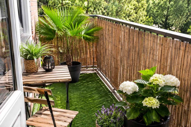 Georganiseerd, licht balkon met bamboe privacyscherm, kunstgras, houten bistroset en diverse potplanten zoals een palm en hortensia.