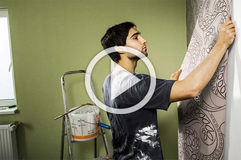 Een man brengt een patroonbehang aan in een kamer, met een ladder en een emmer verf op de achtergrond.