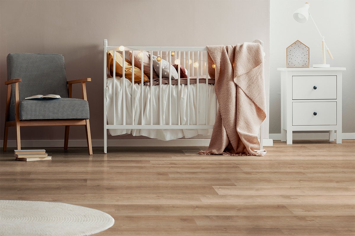 Lichtbruine vloer met houtlook in een lichte babykamer met een wit babybedje, fauteuil met pied-de-poule patroon en wit nachtkastje.
