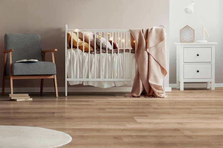 Lichtbruine vloer met houtlook in een lichte babykamer met een wit babybedje, fauteuil met pied-de-poule patroon en wit nachtkastje.