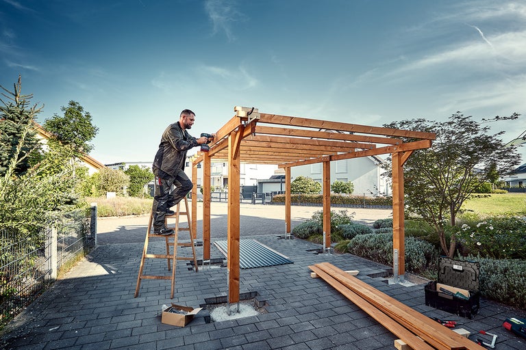 Een man monteert een houten pergola in de tuin met een accuschroevendraaier, staand op een ladder.
