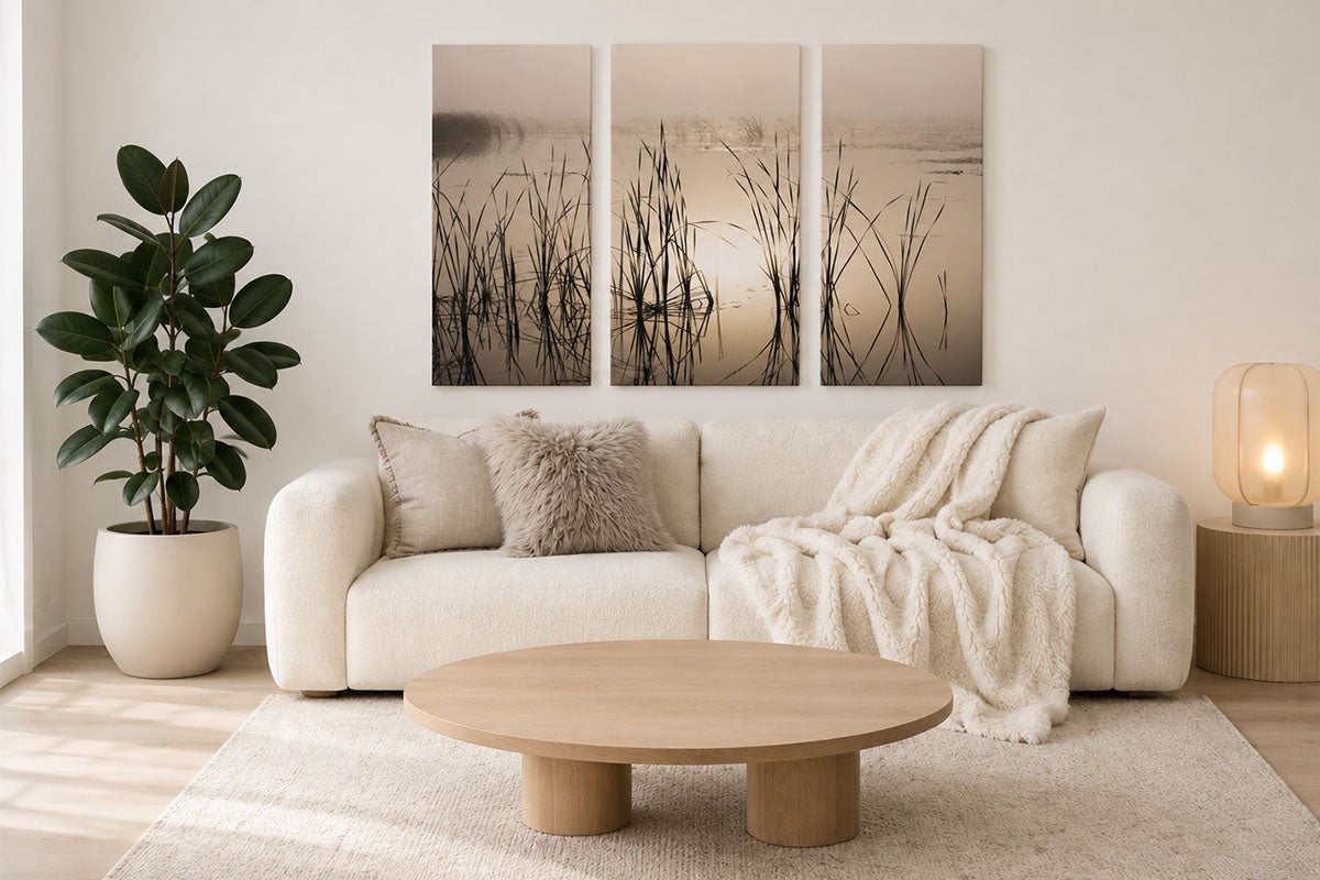 Lichte woonkamer met een witte bank, een ronde houten salontafel en een driedelige wanddecoratie van een mistig landschap met riet.