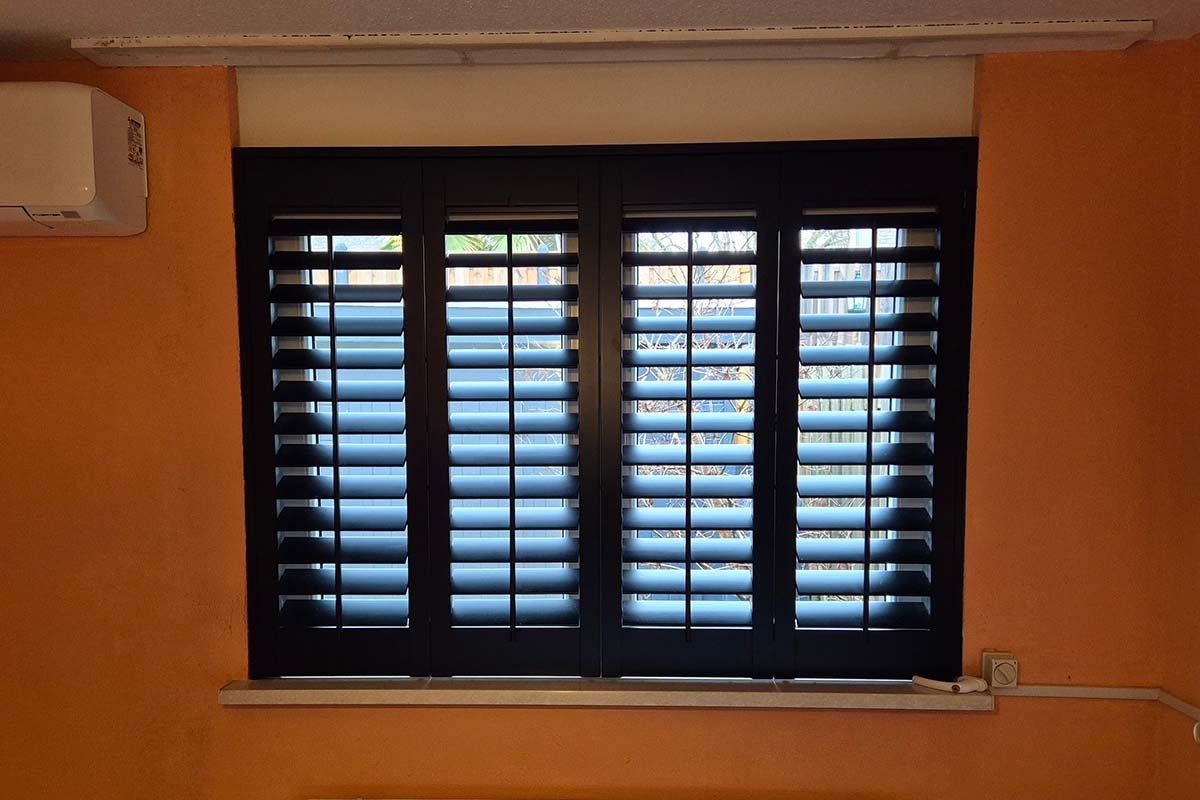 Vierdelige binnenluiken met verstelbare lamellen voor een raam in een kamer met oranje muren.