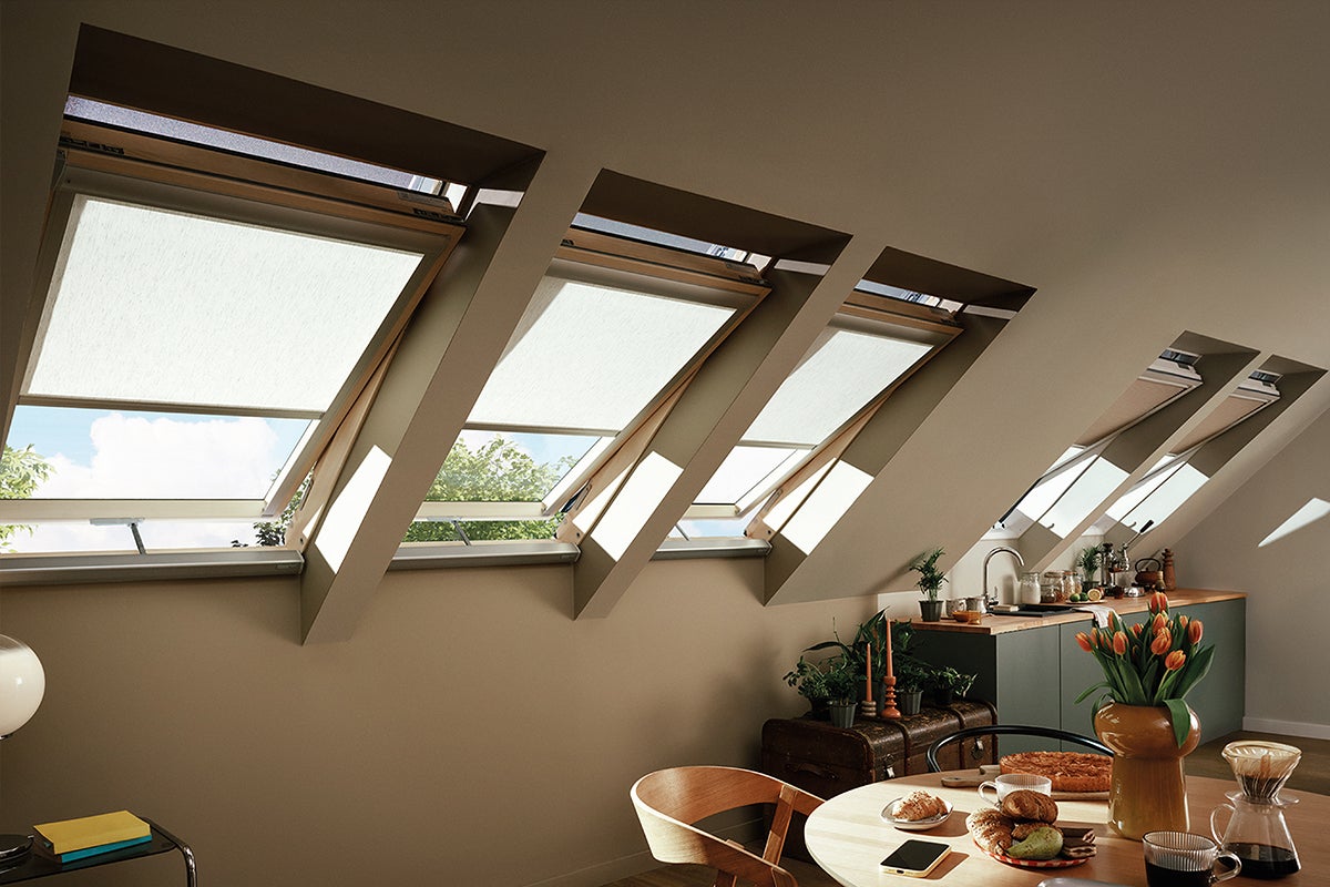 Lichte, moderne zolderkamer met vijf dakramen en rolgordijnen. De ruimte heeft een houten eettafel en een georganiseerd keukenblok.