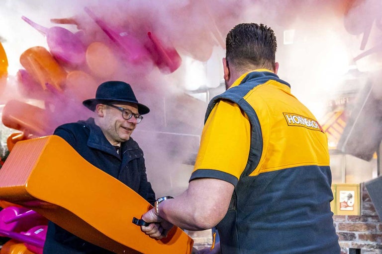 Twee mannen interageren voor een achtergrond van gieters en rook, waarvan er één een werkväst draagt met het Hornbach logo.