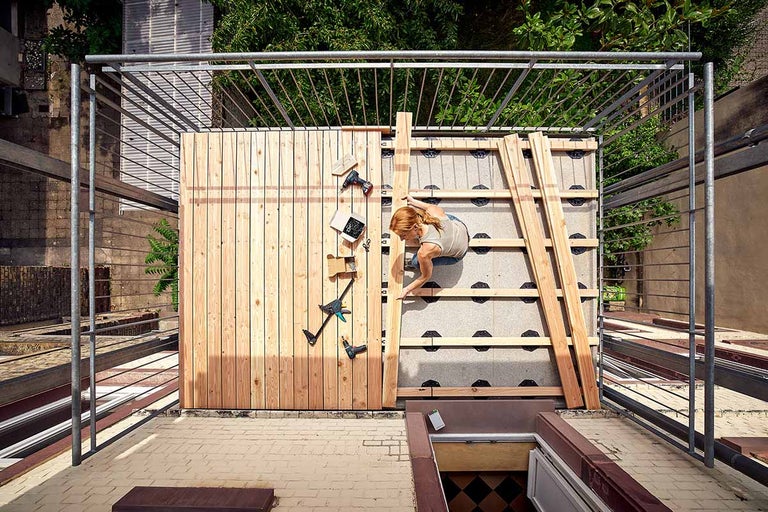Bovenaanzicht van een vrouw die houten vlonderplanken op een balkon met metalen balustrade legt, inclusief gereedschap zoals een accuboormachine.