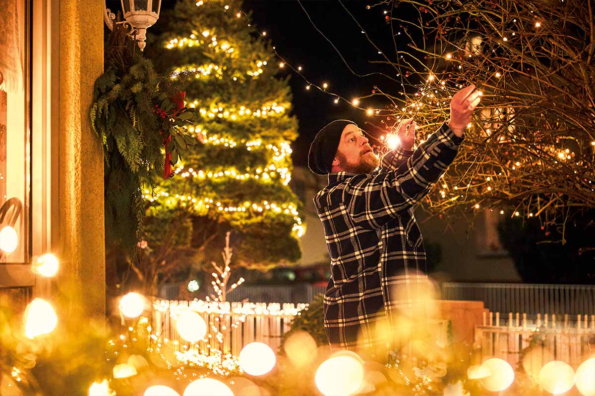 Man versiert een boom met een lichtsnoer buitenshuis. Kerstsfeer met feestelijke verlichting.