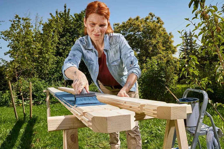 Vrouw schildert houten planken met blauwe houtbeits en een brede kwast in een lichte tuin.