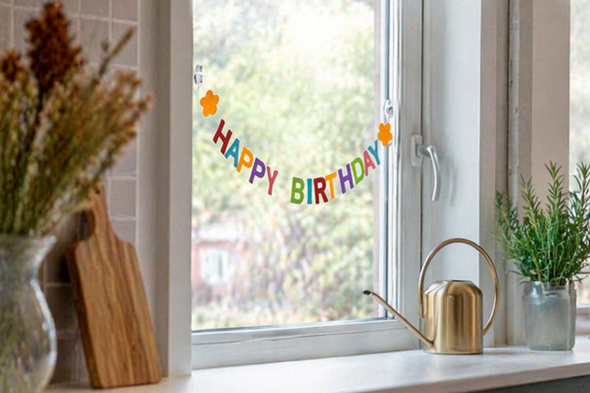 Raamsticker Happy Birthday met gekleurde letters en bloemen op een glazen ruit in een lichte keuken.