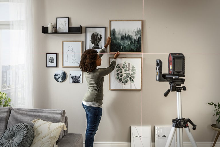 Vrouw gebruikt een laserapparaat om afbeeldingen aan een muur uit te lijnen, met een wandplank met decoratie erboven.