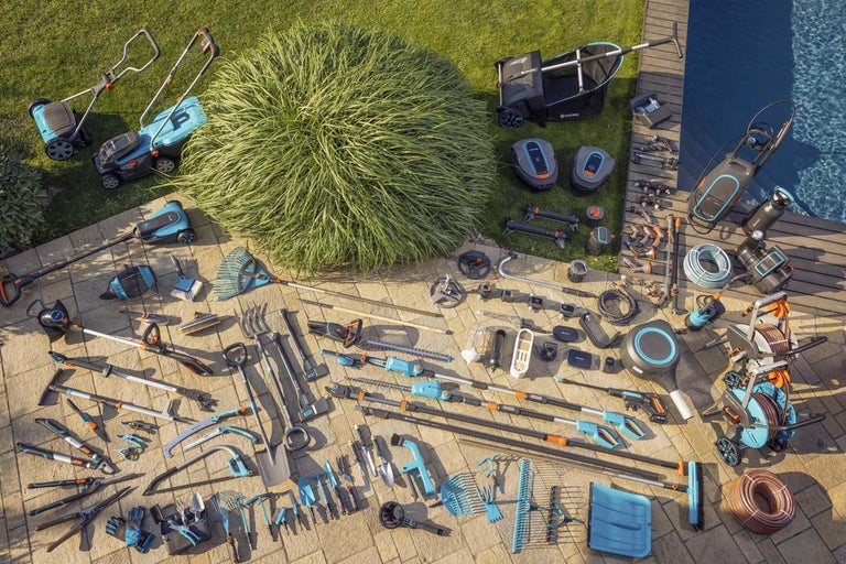 Overzicht van diverse tuingereedschappen op straatstenen in de buitenlucht, waaronder grasmaaiers, robotmaaiers, tuingereedschap en irrigatieproducten