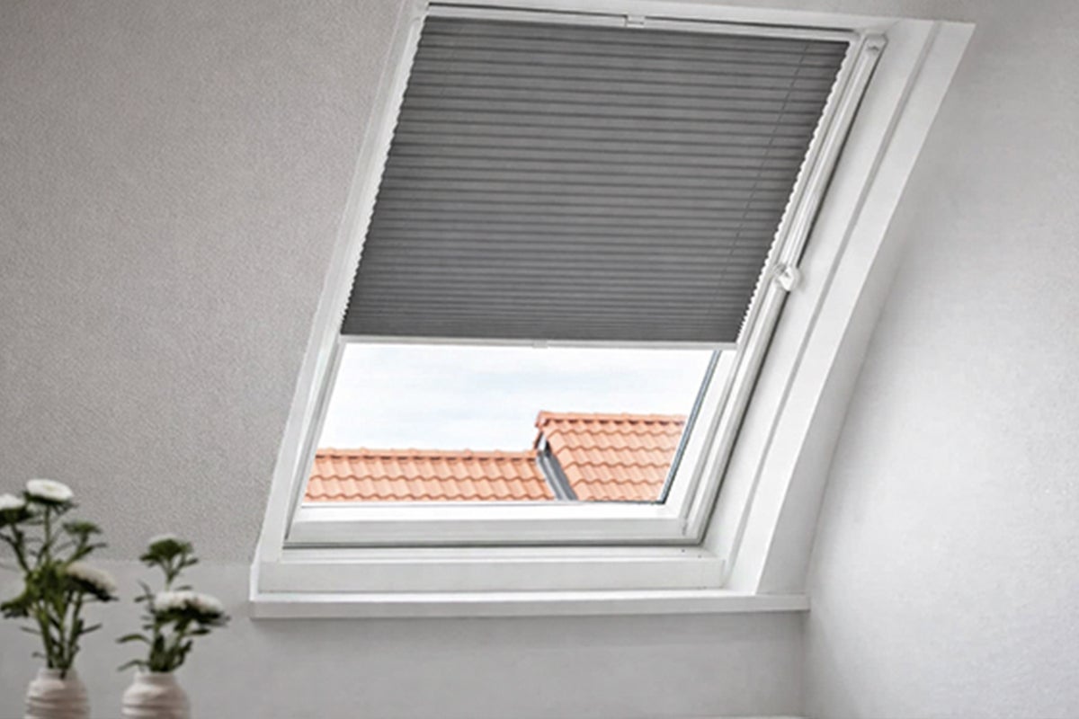 Donker plissé voor dakraam, half gesloten, gemonteerd in een lichte kamer met uitzicht op een pannendak.